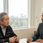 A professional male family lawyer of African descent is in a consultation with an elderly Asian male client in a bright Mississauga office. They are seated at a wooden desk with visible legal documents and a city skyline including the Absolute World towers in the background.